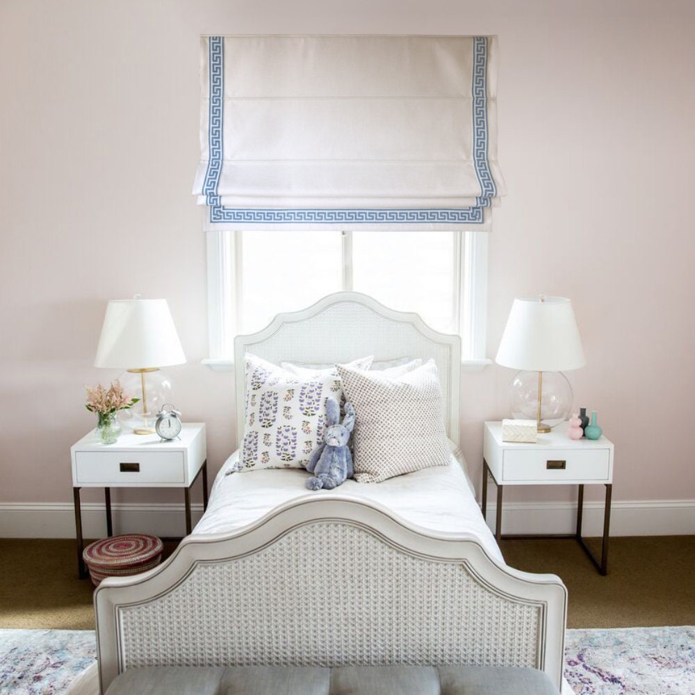 White flat Roman shade with blue Greek key trim in a pale pink bedroom with a white wicker bed