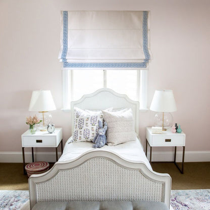 White flat Roman shade with blue Greek key trim in a pale pink bedroom with a white wicker bed