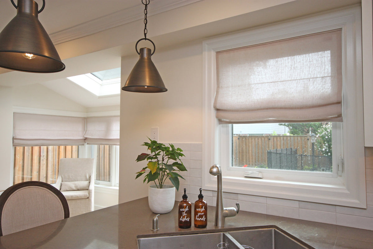Custom Zoe Italian linen relaxed Roman shade over a kitchen sink, soft folds, pendant lights and a plant