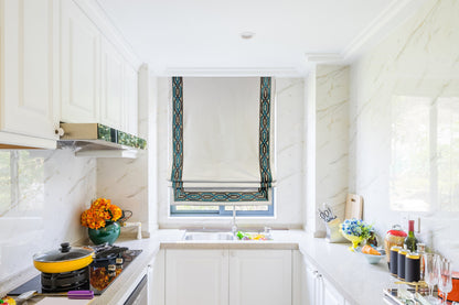 Flat Roman shade in white linen with teal and gold woven trim, shown in a bright room