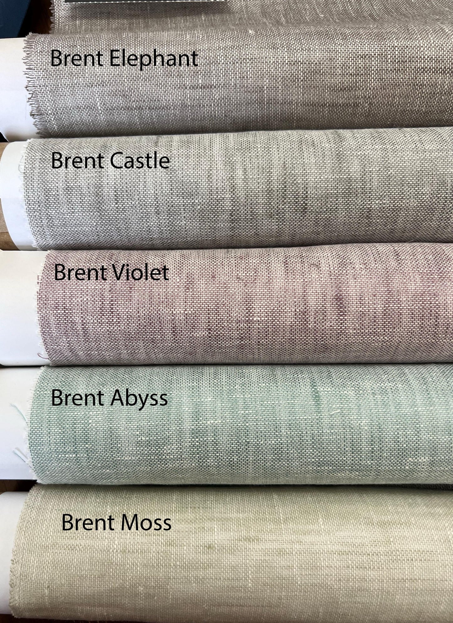 Rolled sheer linen fabrics stacked and labeled Brent Elephant, Brent Castle, Brent Violet, Brent Abyss and Brent Moss