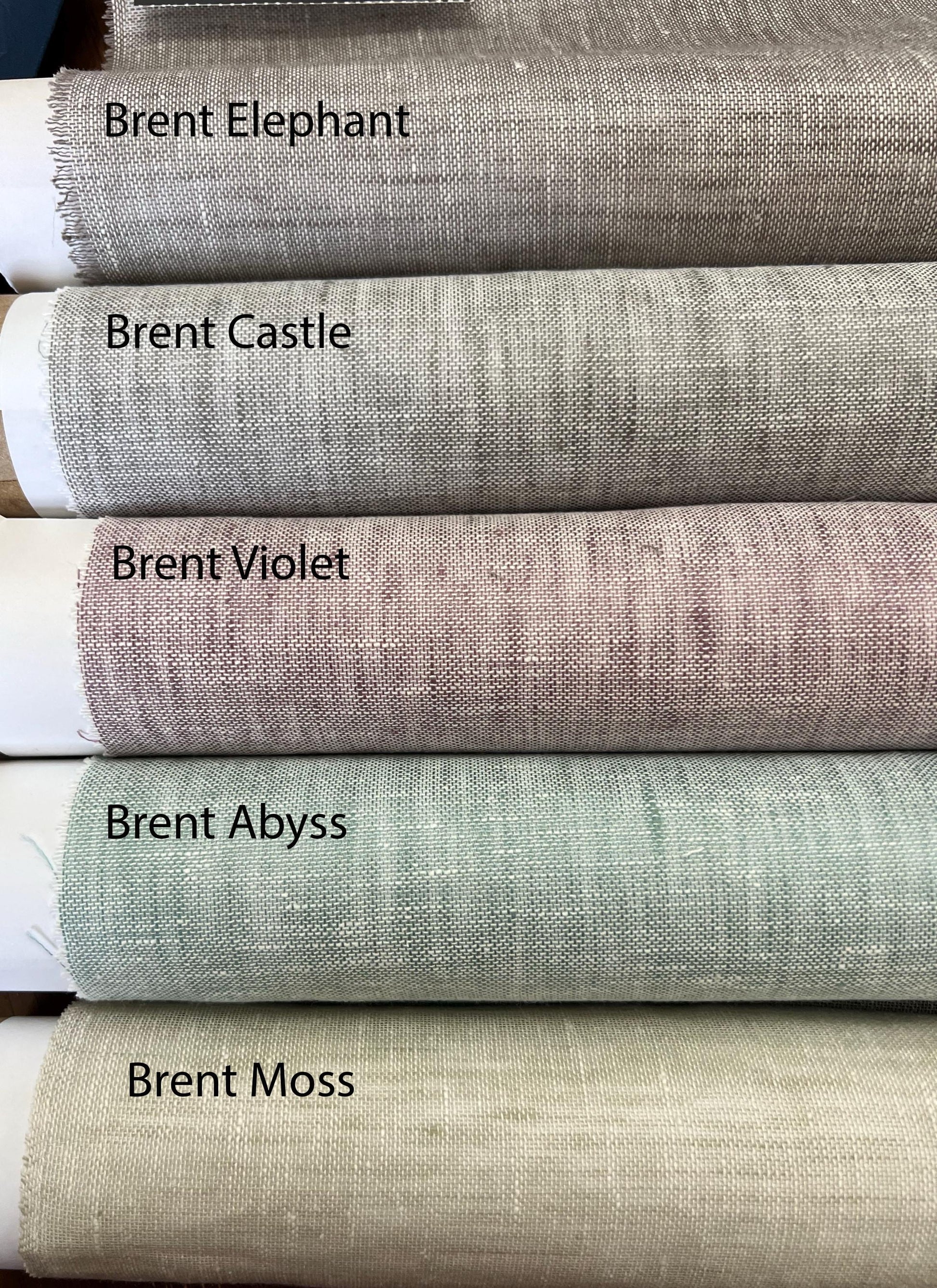 Rolled sheer linen fabrics stacked and labeled Brent Elephant, Brent Castle, Brent Violet, Brent Abyss and Brent Moss