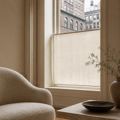 Ivory boucle armchair beside a window with a sheer linen cafe curtain filtering light from brick city buildings