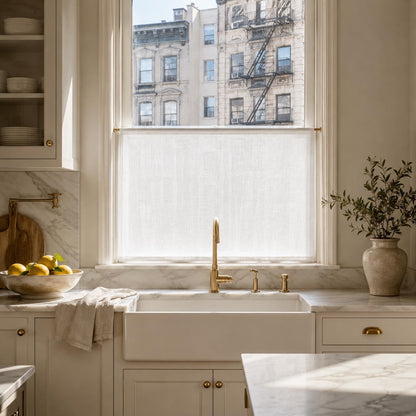 White sheer linen cafe curtain covering the lower half of a kitchen window above a farmhouse sink, with brass faucet, marble counters and a city view