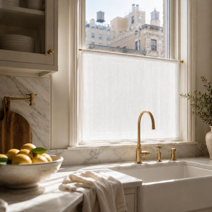 Custom sheer cafe curtain on the lower sash of a kitchen window, with marble backsplash, brass faucet, bowl of lemons and historic buildings outside