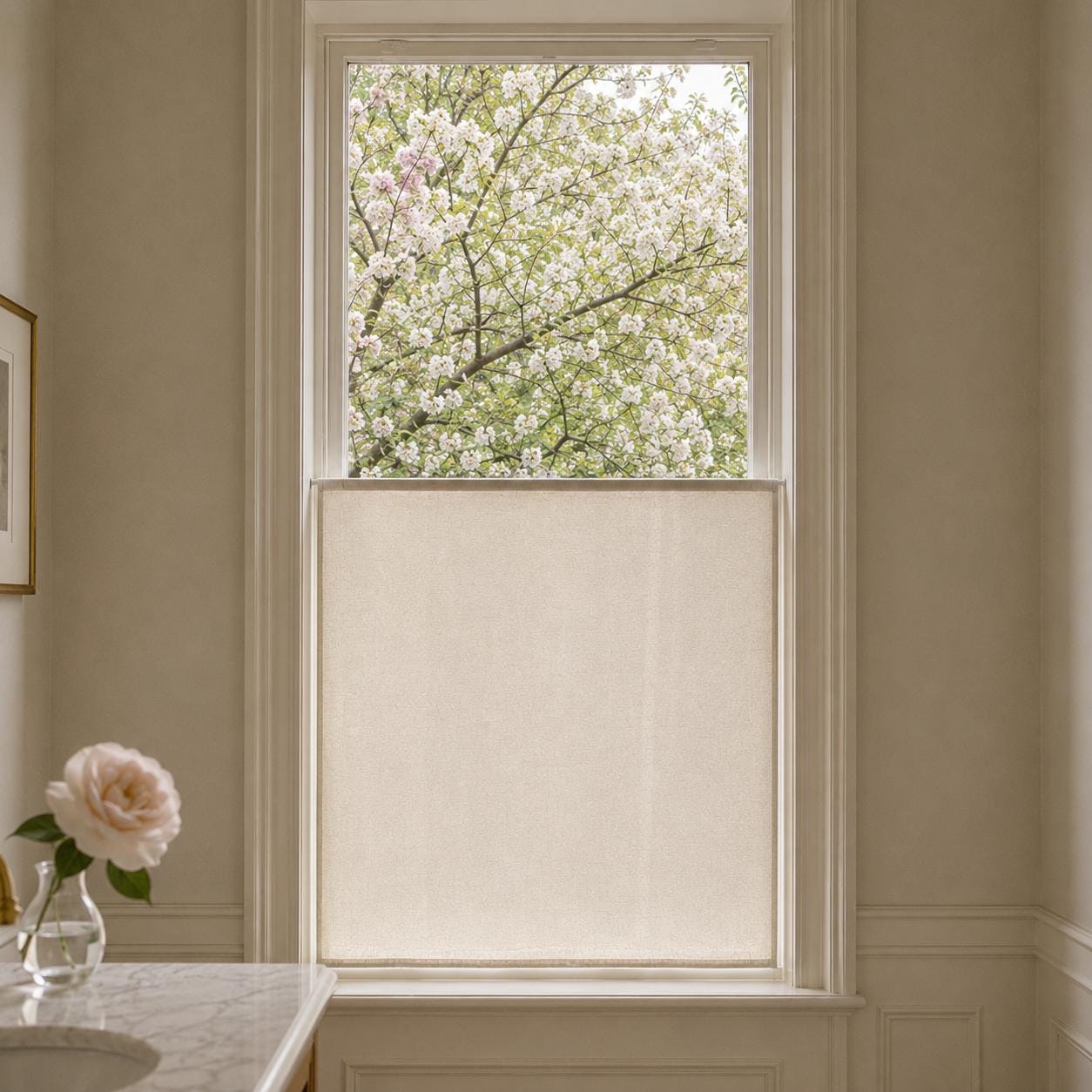 Flat sheer cafe curtain mounted on the lower sash of a tall window, a rose in a vase on the marble ledge and blossoming trees outside