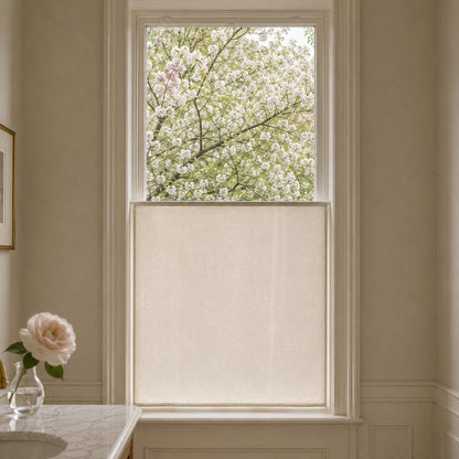 Flat sheer cafe curtain mounted on the lower sash of a tall window, a rose in a vase on the marble ledge and blossoming trees outside