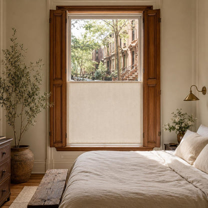Bedroom with linen bedding and a wood-framed window, sheer cafe curtain covering the lower half with brownstones outside
