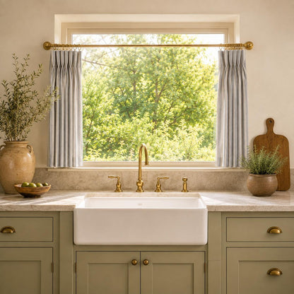 Grey striped cafe curtain above a white farmhouse sink with a brass bridge tap and greenery.