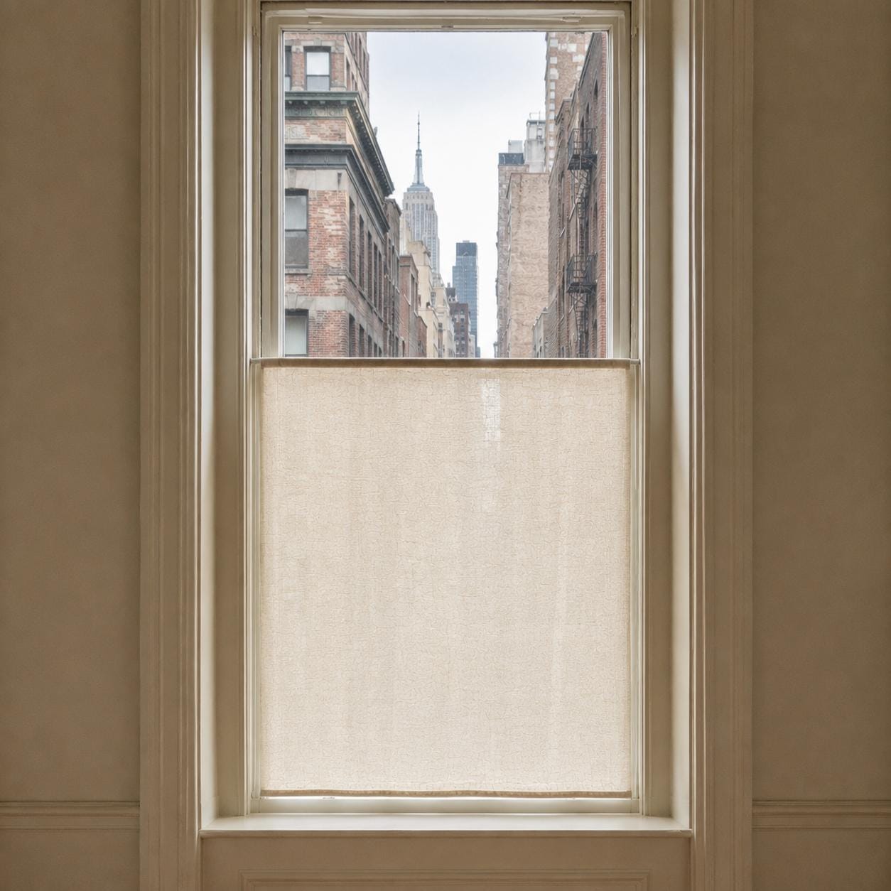 Tall city window with a flat sheer cafe curtain on the lower half, overlooking a New York street view