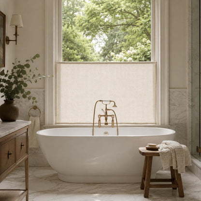 Sheer cafe curtain screening the lower window behind a freestanding white bathtub, with brass tub filler and greenery outside