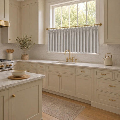 Pinch pleated cafe curtain at a kitchen window above a marble counter in a cream kitchen.