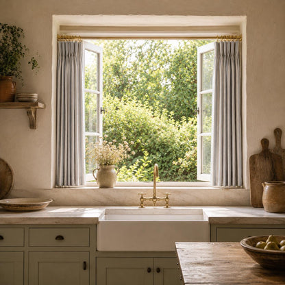Open casement window over a farmhouse sink framed by grey ticking stripe cotton curtains.