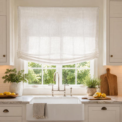 Sheer white linen relaxed Roman shade above a farmhouse kitchen sink in a white kitchen