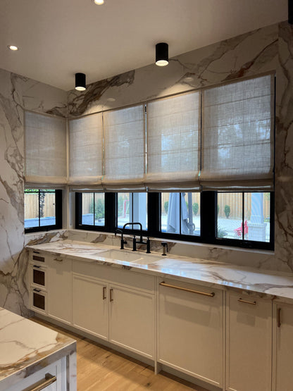 Linen Roman shades across black framed windows in a white marble kitchen