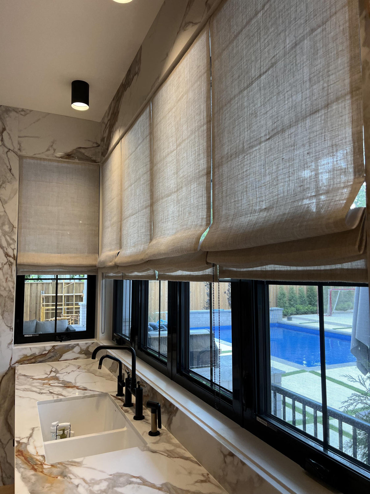 Linen Roman shades along a wall of windows in a marble kitchen looking out to a pool