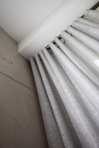 Grey Brent sheer ripple fold drapery on a ceiling track against a concrete wall, viewed from below